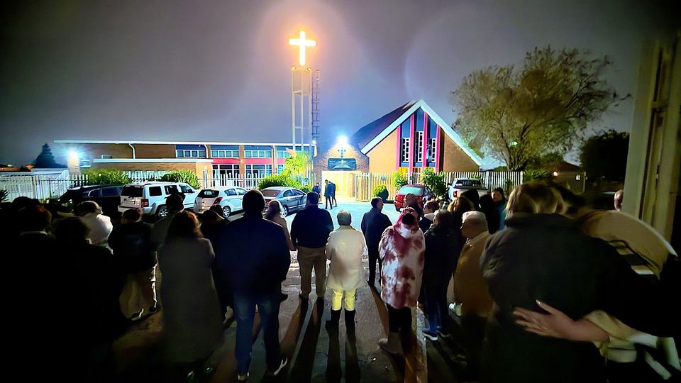 Die gemeente saans buite die kerk onder die verligte kruis op die toring